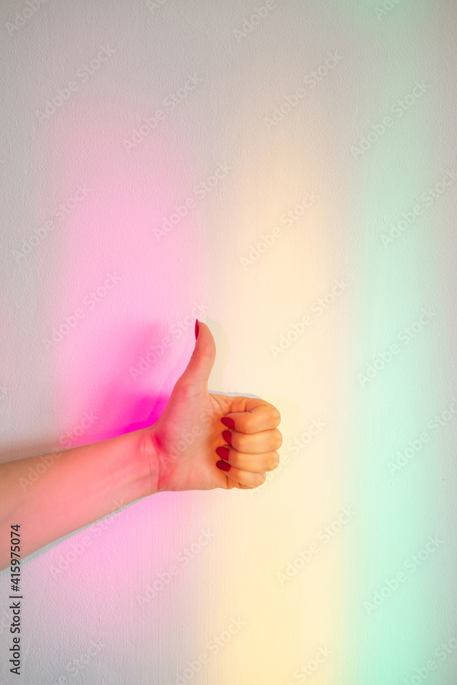 Like gesture. Approving sign. Good job. Favor support. Yes symbol. Female hand with thumb up on blur rainbow glowing neon lights wall background. Like gesture. Approving sign. Good job. Favor support. Yes symbol. Female hand with thumb up on blur rainbow glowing neon lights wall background.