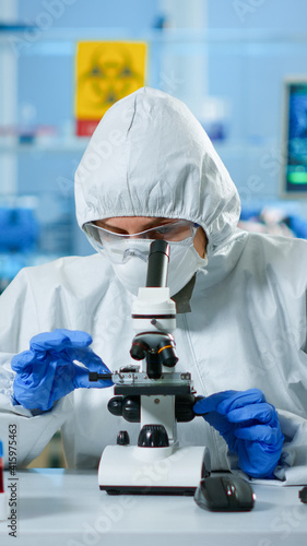 Scientist in ppe suit making adjustments and looking through laboratory microscope, typing on pc. Chemist in coverall working with various bacteria, tissue blood samples for antibiotics research