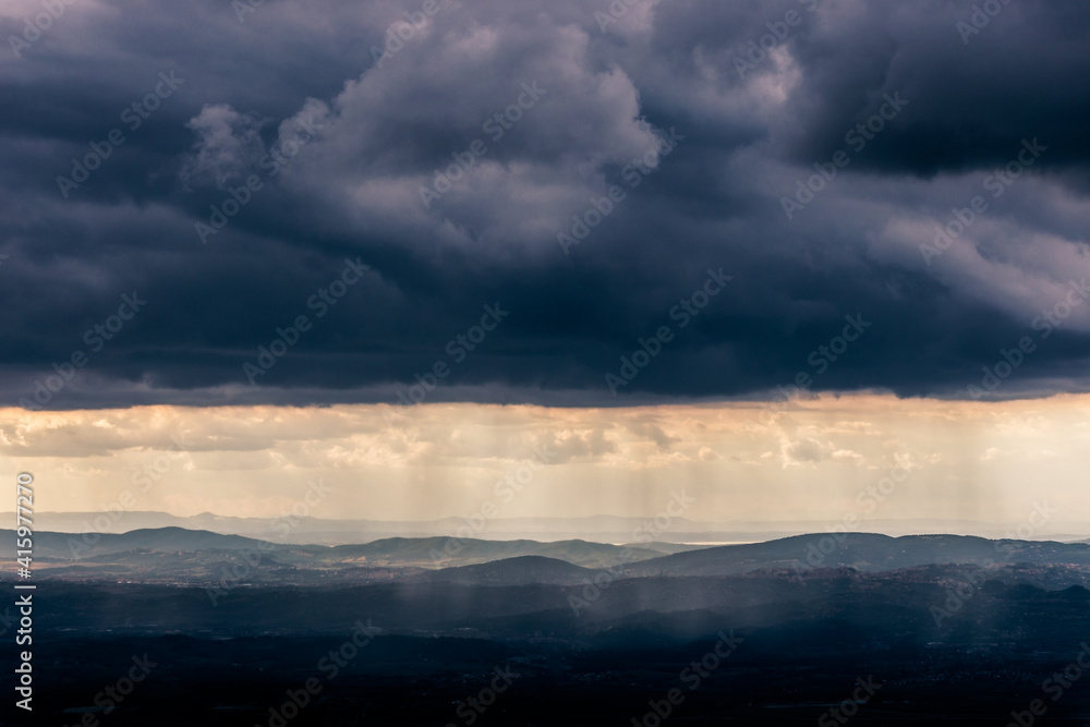 Mist and sunrays between valley and layers of mountains and hills beneath a moody, overcast sky with heavy clouds