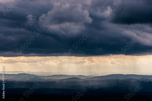 Mist and sunrays between valley and layers of mountains and hills beneath a moody, overcast sky with heavy clouds