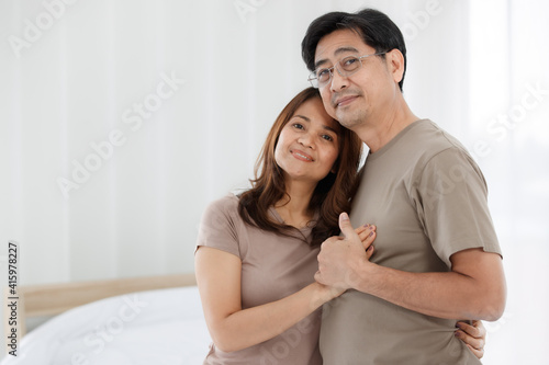 Half-body horizontal portrait shot of cute smiling senior Asian lover couple standing at home. Husband and wife holding hands together, touching at man heart and looking at each other eyes.