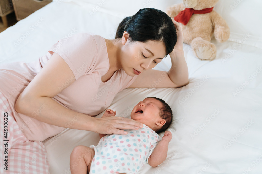 chinese mother feeling heart ached with pity looking at her baby crying out of illness. new mother propping head lying bedside patting on her newborn girl’s chest to coax her...