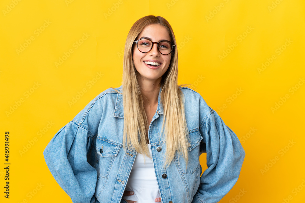 Young blonde woman isolated on yellow background posing with arms at hip and smiling