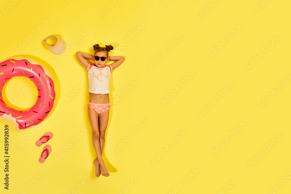 Fototapeta premium little child girl in swimwear and sunglasses lying on yellow background with inflatable rubber ring. Top view. copy space