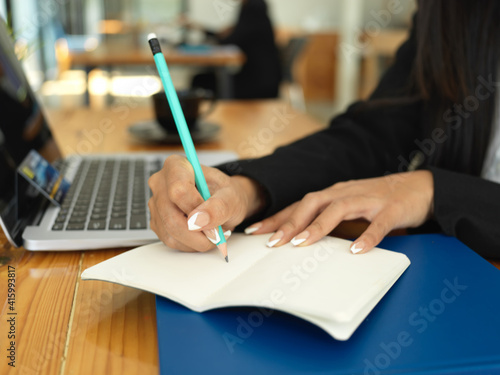 Businesswoman hand with pencil writing on notebook while working with laptop