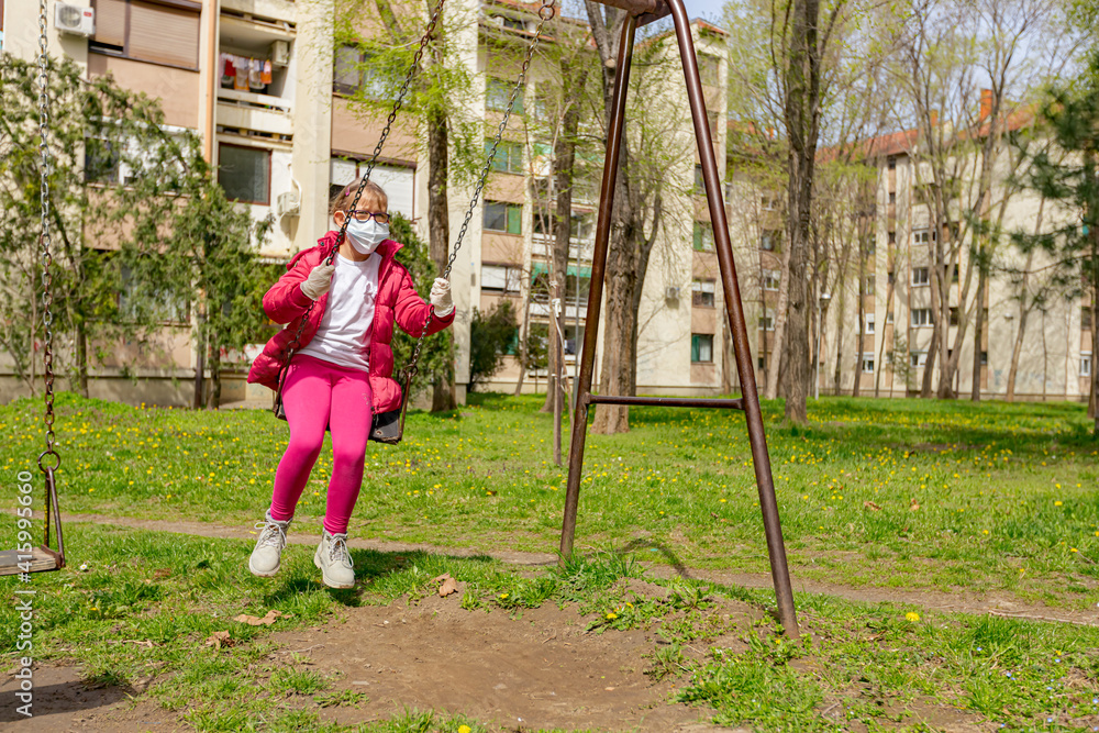 Little girl in medical mask and gloves is swinging alone.