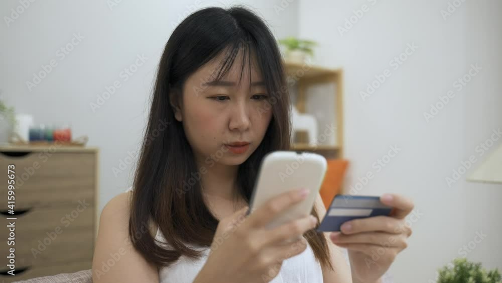 portrait attractive asian girl holding phone is paying online with her credit card and entering numbers carefully while having fun shopping on the internet at home. portrait attractive asian girl holding phone is paying online with her credit card and entering numbers carefully while having fun shopping on the internet at home.