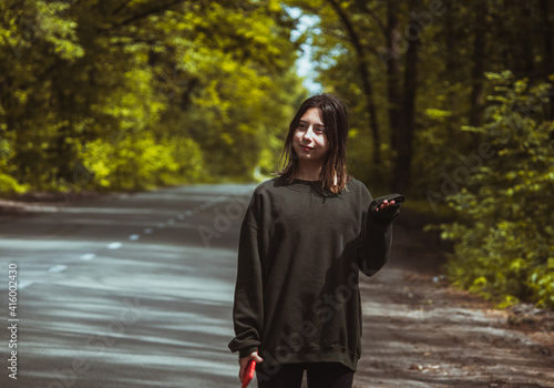 A girl in a large green sweatshirt stands with a sarcastic surprised face in the middle of a forest trail on a sunny day.