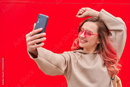 Young blogger woman with pink dyed hairs staying near the red wall and making selfie by smartphone