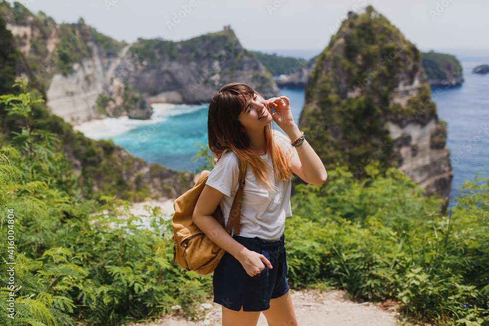 Naklejka premium Traveling and adventure concept. Happy pretty woman with back pack traveling in Indonesia on Nusa Penida island. Amazing cliffs and tropical beach view on background.