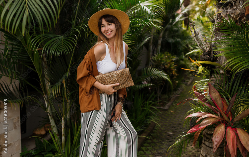Tropical vacation and holidays concept.  Elegant stylish girl in white top and straw hat posing on palm leaves background in Bali.