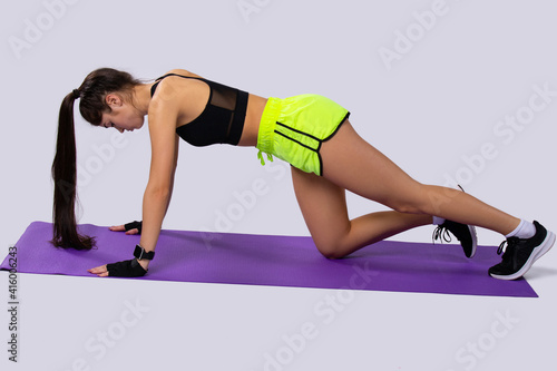 Fototapeta Naklejka Na Ścianę i Meble -  Side view of slim, pretty, positive, young brunette woman doing exercise, on violet sport mat, on gray background. Yoga, sport and pilates