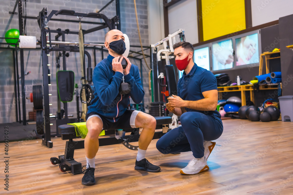 Instructor in the gym correcting the squats of the young athlete in the coronavirus pandemic, a new normal. With protective face mask