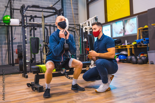 Instructor in the gym correcting the squats of the young athlete in the coronavirus pandemic  a new normal. With protective face mask