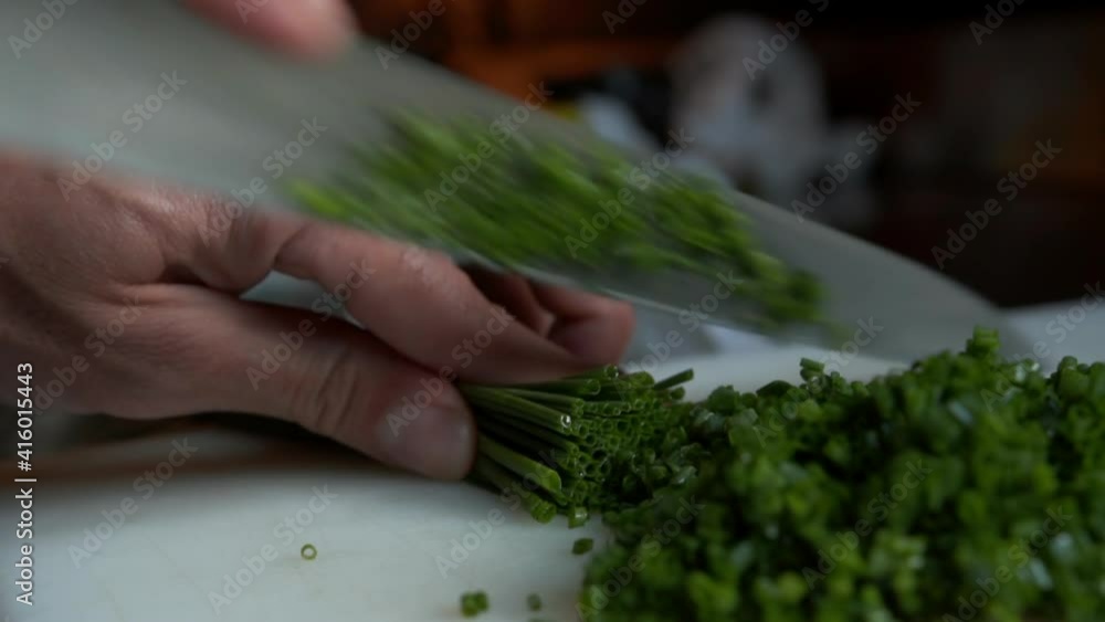 Close up view of knife cutting multiple chives into pile of small vegetable pieces. Preparing green vegetables on the kitchen table Close up view of knife cutting multiple chives into pile of small vegetable pieces. Preparing green vegetables on the kitchen table
