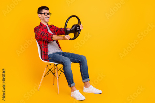 Photo of funny man wear checkered shirt eyewear sitting chair holding steering wheel empty space isolated yellow color background