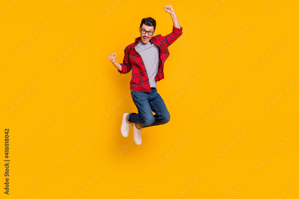 Full length photo of lucky young guy dressed plaid shirt eyeglasses jumping high rising fists isolated yellow color background
