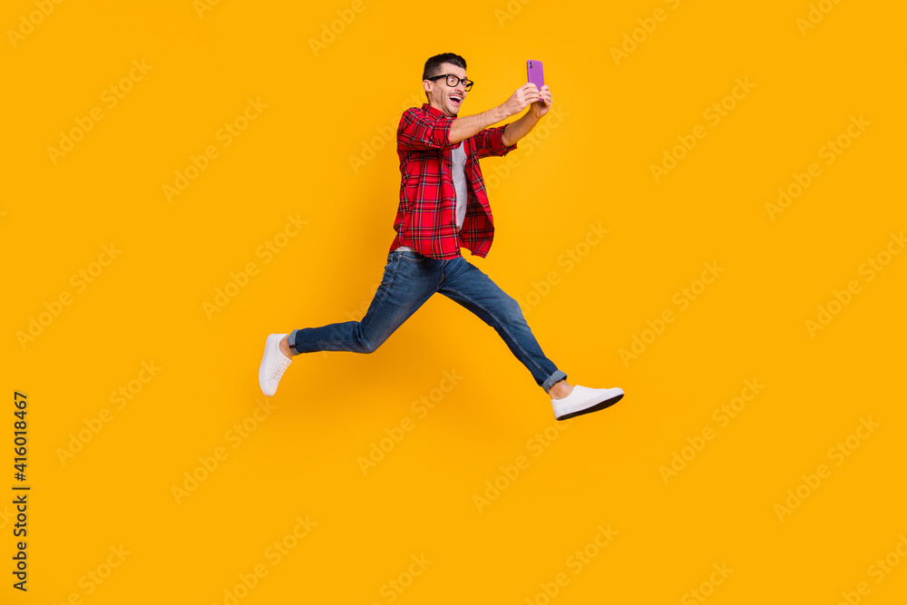 Full length photo of cute young guy dressed plaid shirt eyeglasses jumping holding modern device isolated yellow color background