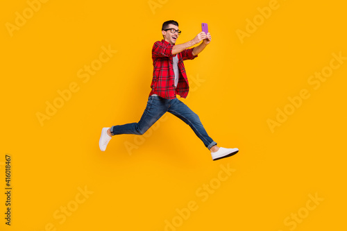 Full length photo of cute young guy dressed plaid shirt eyeglasses jumping holding modern device isolated yellow color background