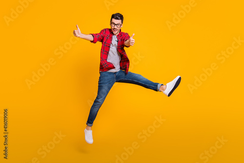 Full body photo of brunette man make thumbs up jump air wear jeans glasses isolated on yellow color background