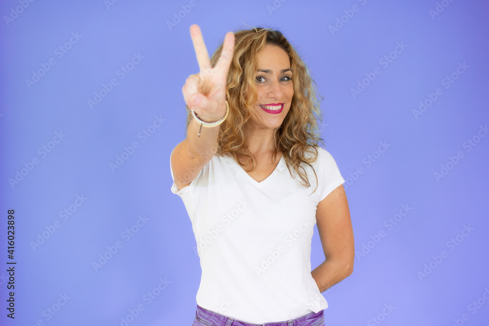Fototapeta premium Blonde woman with curly hair wearing casual t-shirt smiling with happy face winking at the camera doing victory sign. number two.