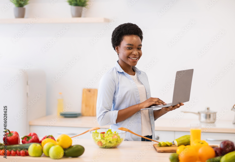 African Lady Holding Laptop Browsing Recipes Cooking Dinner In Kitchen African Lady Holding Laptop Browsing Recipes Cooking Dinner In Kitchen