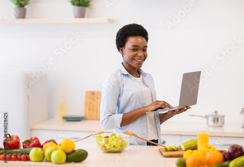 African Lady Holding Laptop Browsing Recipes Cooking Dinner In Kitchen