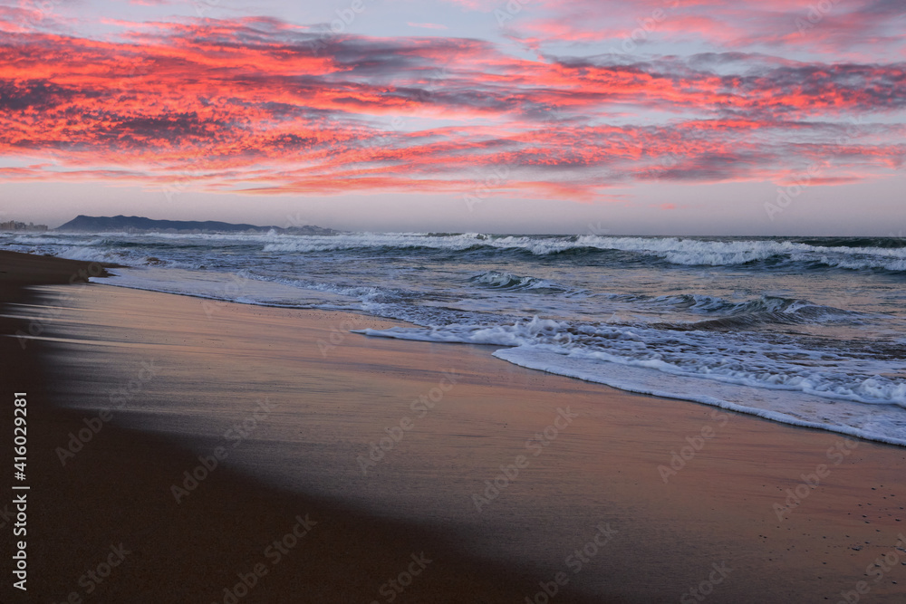 Beautiful sunset with red clouds and waves on a beach in Valencia, Spain