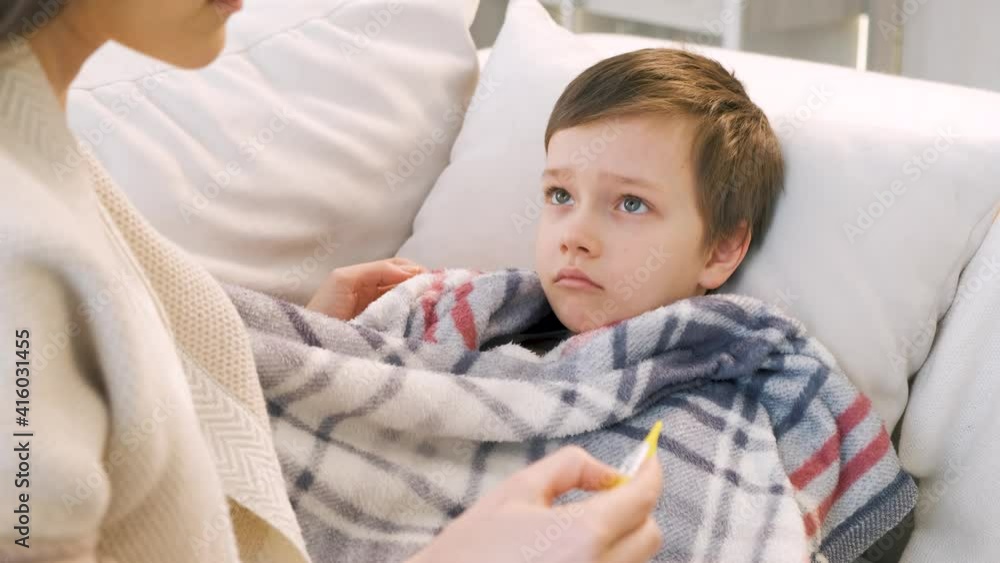 Sick little boy covered in blanket is lying on couch while his mother is taking his temperature Sick little boy covered in blanket is lying on couch while his mother is taking his temperature
