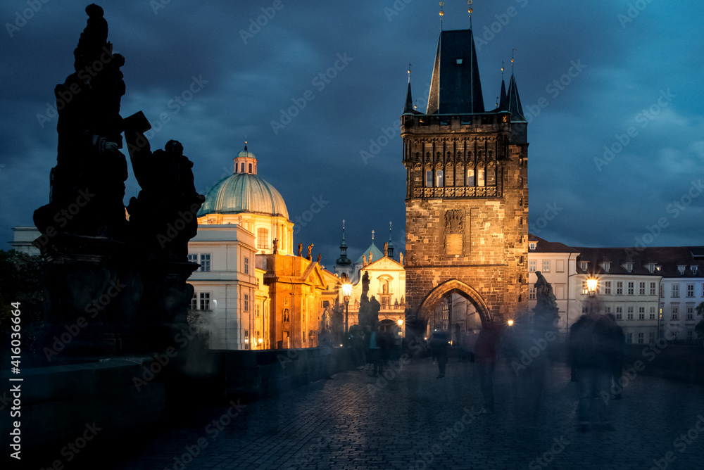 Blurred ghost like people silhouettes walking on illuminated Charles Bridge in Prague in night on Gothic towers background