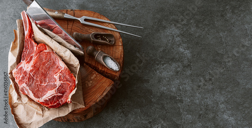 Photos on a wooden block for meat a fresh raw tamahawk steak or a cowboy steak with a butcher's chopping ax for meat, next to it is a mixture of peppers and coarse salt, milled with thyme