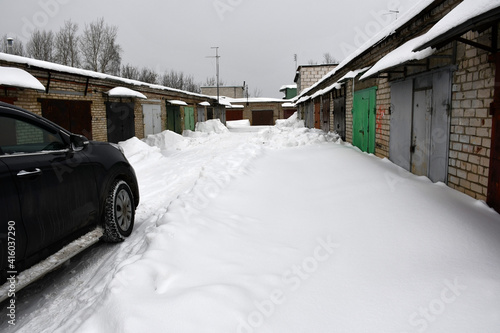 A cloudy winter day. Snowfall. The car is parked between one-story brick garages with closed metal painted gates. Drifts of snow near the walls along the road. Snow on the roofs. © Olga