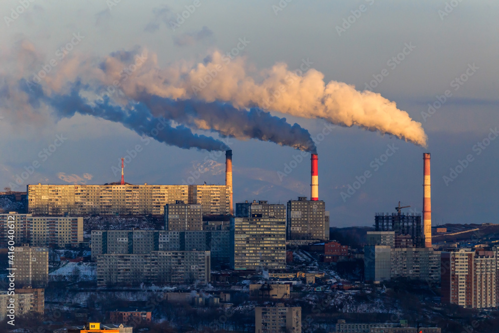 The smoking chimneys of TPP-2 in Vladivostok amid residential buildings and distant snowy mountains. Smoke in the city.