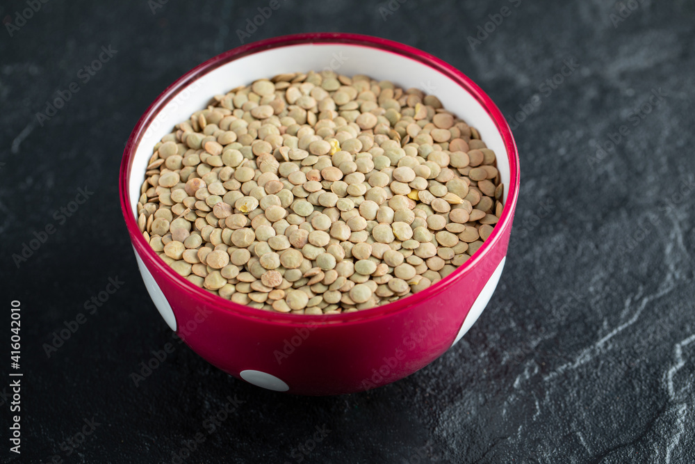 A red bowl with dried brown raw lentils on a dark background A red bowl with dried brown raw lentils on a dark background