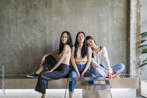 Three happy young Asian female friends casually pose sitting together inside a rustic room against bare concrete wall © kudosstudio