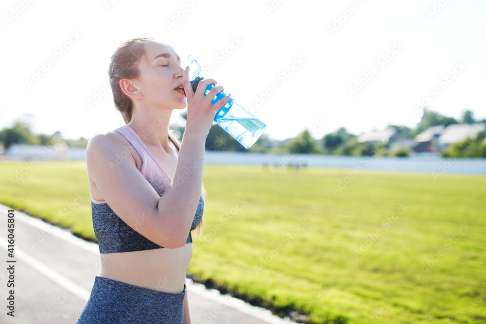 Slim young girl trains in the stadium and holds a bottle of water. The girl is training outdoors Slim young girl trains in the stadium and holds a bottle of water. The girl is training outdoors