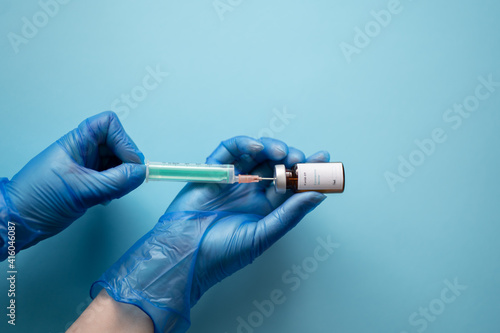 A nurse wearing blue gloves, holding a vial of coronavirus vaccine with one hand, and with the other a needle, with a blue background