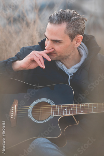 A young white man, with gray hair, musician and artist, plays an elegant black guitar, and composes music outside, in the countryside, on a cloudy afternoon. photo