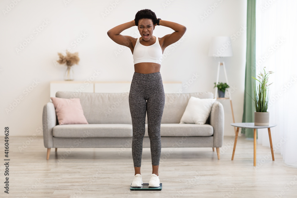 Discontented African American Woman Shouting Standing On Weight-Scales At Home Discontented African American Woman Shouting Standing On Weight-Scales At Home