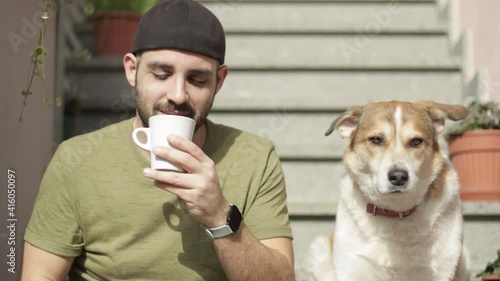 Caucasian man drinks a cup of tea outdoor for break or breakfast not to his brown and white or red dog