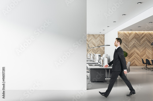 Businessman in black suit goes by modern workplaces in sunny open space office with blank light wall. Mock up © Who is Danny