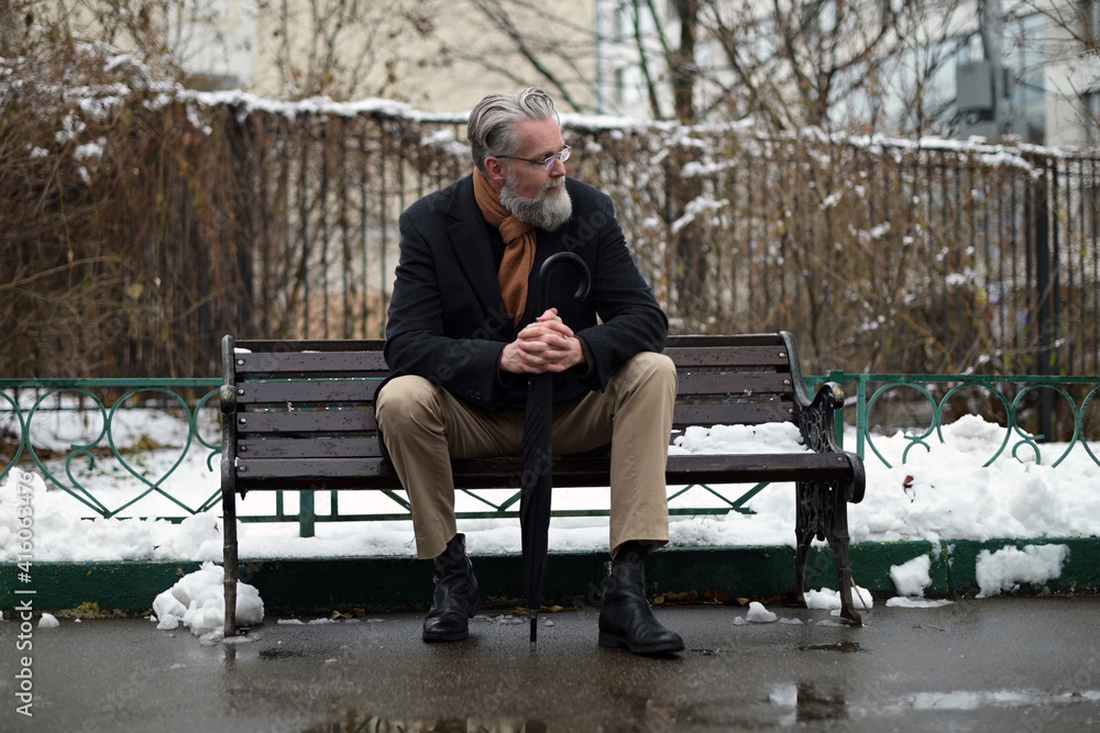 An adult gray-haired respectable man with glasses sits on a park bench and enjoys a winter morning