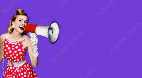Beauty blond haired woman holding megaphone, shout something. Girl in red pin up style dress in polka dot, isolated over purple violet background. Caucasian model in retro and vintage studio concept.