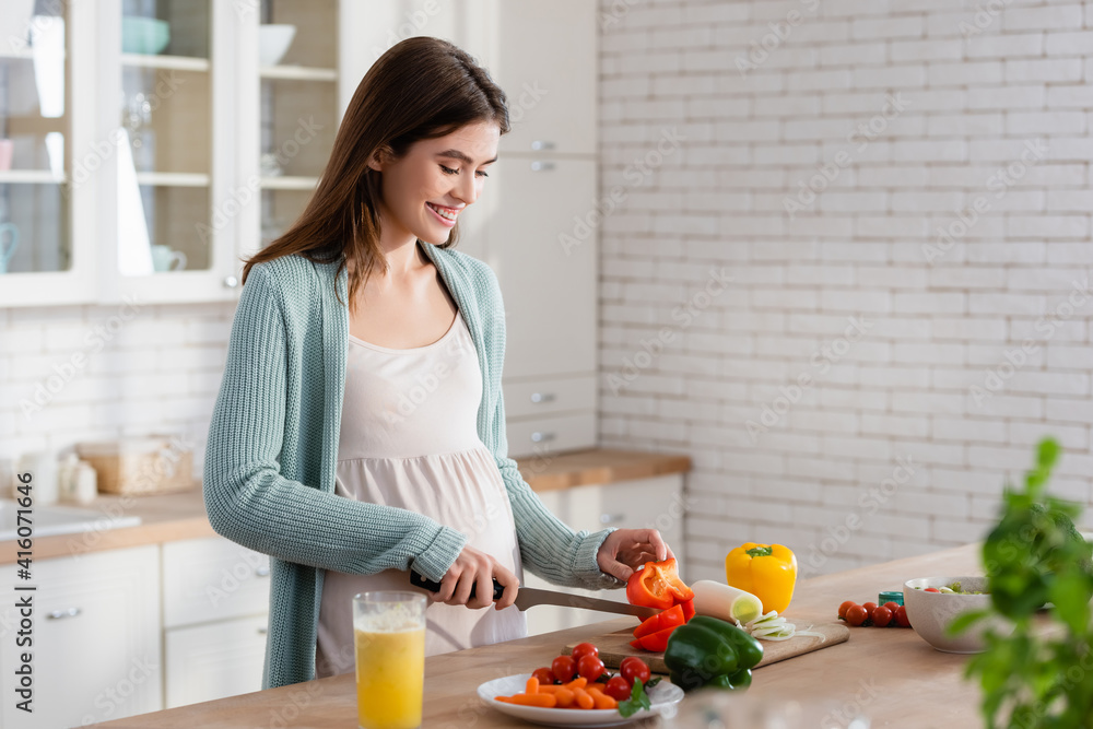 smiling pregnant woman cutting fresh vegetables in kitchen