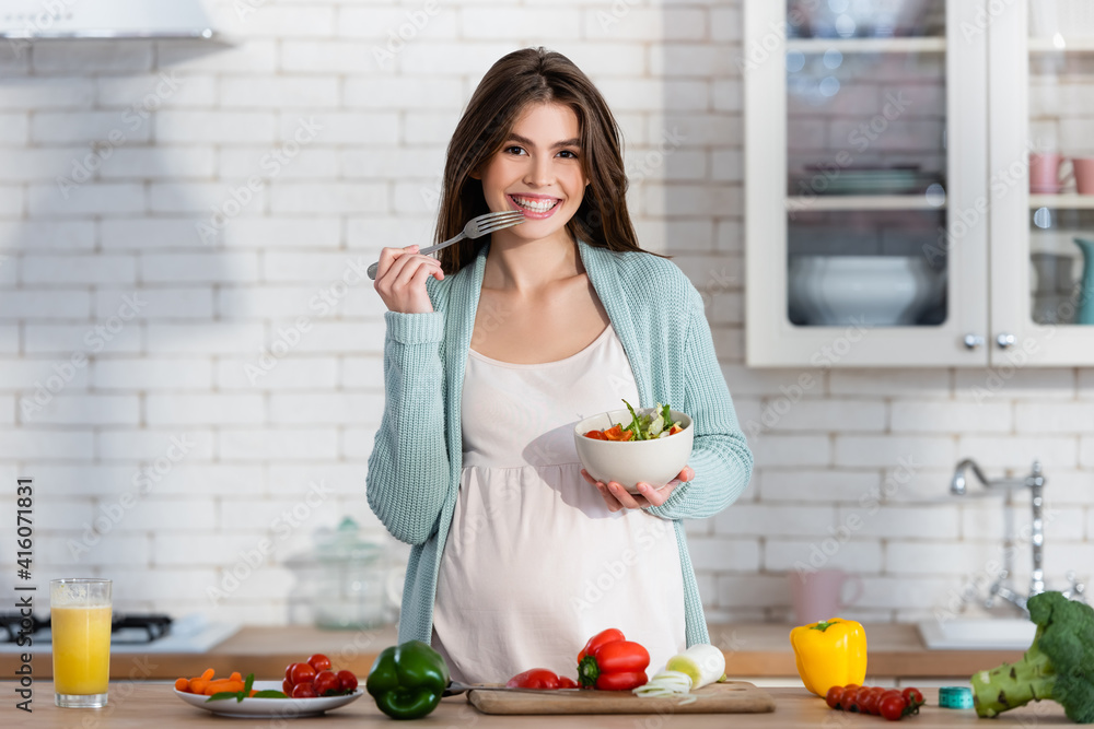 cheerful pregnant woman eating salad near fresh vegetables in kitchen cheerful pregnant woman eating salad near fresh vegetables in kitchen