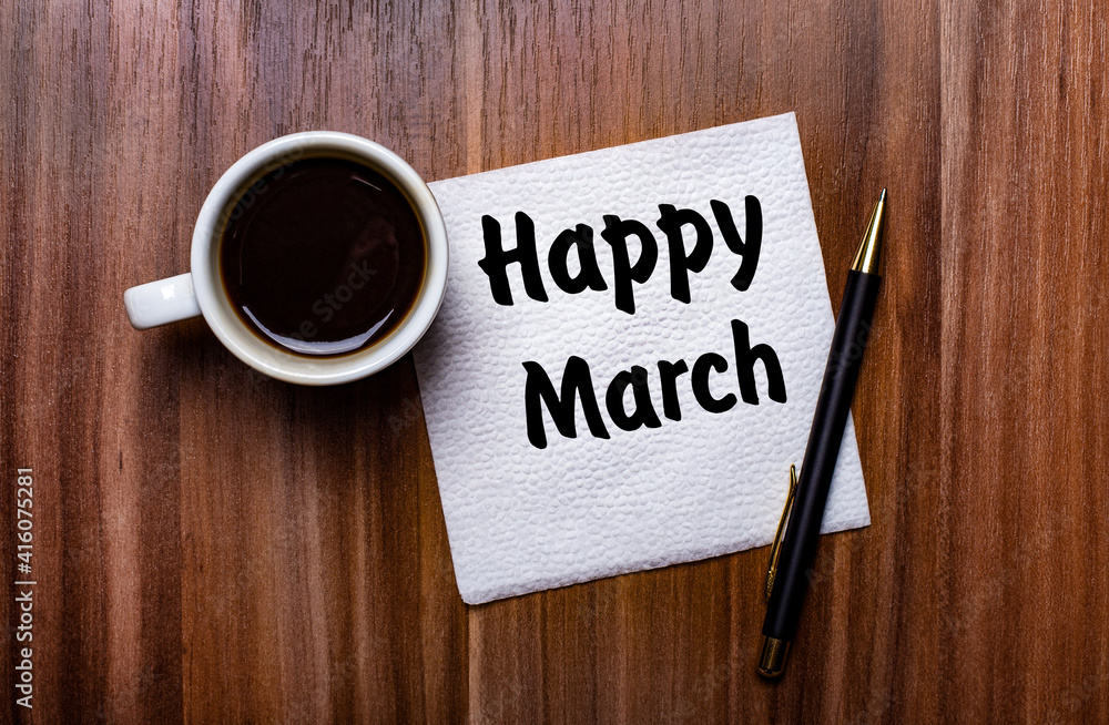 On a wooden table next to a white cup of coffee and a pen is a white paper napkin with the words HAPPY MARCH On a wooden table next to a white cup of coffee and a pen is a white paper napkin with the words HAPPY MARCH