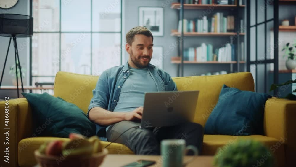 Handsome Caucasian Man Working on Laptop Computer while Sitting on a Sofa Couch in Stylish Cozy Living Room. Freelancer Working From Home. Browsing Internet, Using Social Networks, Having Fun. Handsome Caucasian Man Working on Laptop Computer while Sitting on a Sofa Couch in Stylish Cozy Living Room. Freelancer Working From Home. Browsing Internet, Using Social Networks, Having Fun.