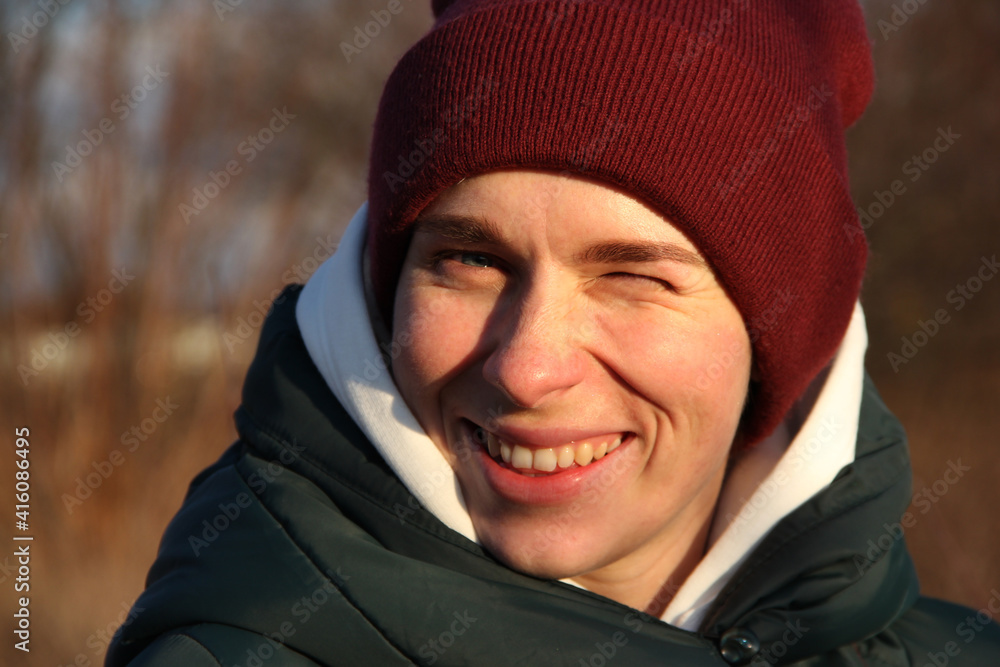 A blue-eyed girl in a burgundy knitted hat and a white hood smiles, winking with one eye against the background of a spring forest.
A blue-eyed girl in a burgundy knitted hat and a white hood smiles, winking with one eye against the background of a spring forest.