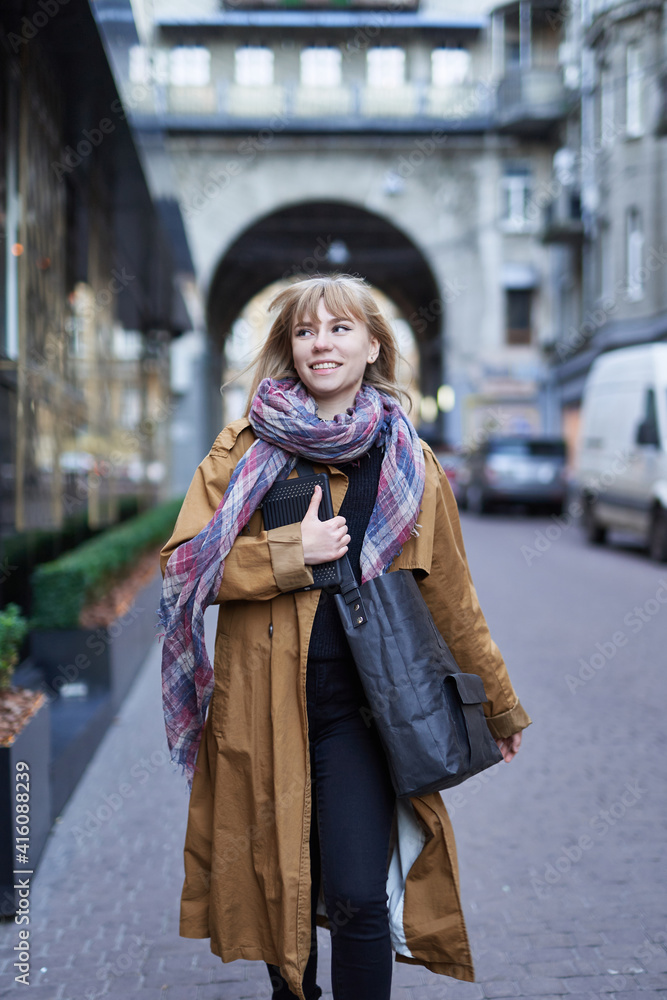 custom made wallpaper toronto digitalStreet style photo of elegant blonde woman wearing long orange trench coat, black jeans and scarf with black zero waste bag, walking on street of European city
