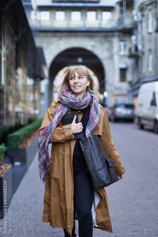 Street style photo of elegant blonde woman wearing long orange trench coat, black jeans and scarf with black zero waste bag, walking on street of European city